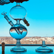 Load image into Gallery viewer, Glass water pipe with blue liquid on a reflective surface, blurred cityscape in the background
